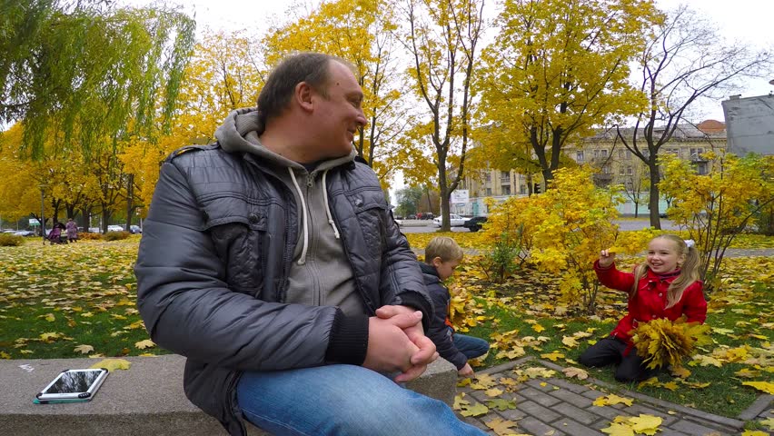 happy family playing with autumn leaves in park. Boy and girl collect yellow leaves in the park. Father sitting nearby.