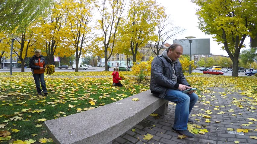 Father sitting on a park bench and read e-books. The children gather leaves. Yellowed trees in autumn.