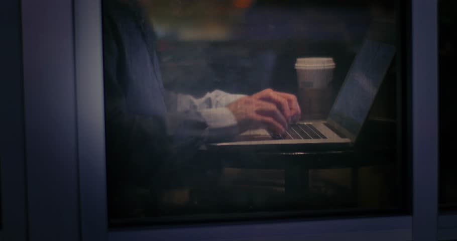 Businessman working late on laptop computer while in cafe shop, closeup on hands typing on keyboard. View through window, shallow DOF.