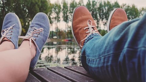 Point Of View: Couple sitting on a wooden jetty playing footsie, close up on modern hipster shoes. 4K Ultra HD. Relaxed time by the lake on a pier. POV: romantic young love by the lakeside. Fashion.