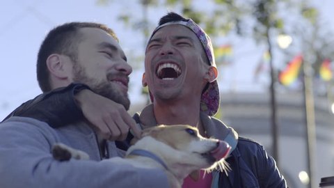 Cute Couple Hold Their Jack Russell Terrier And Get Dog Kisses, Men Kiss Each Other, Gay Pride Flags Blow In Breeze Behind Them Video de stock