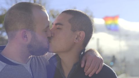Portrait Of Cute Male Couple, They Kiss, Gay Pride Flag Blows In Breeze Behind Them