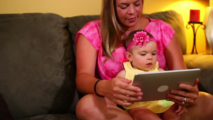 A mother uses a tablet computer with her young baby on the sofa.