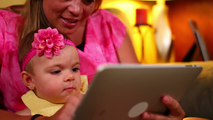 A mother uses a tablet computer with her young baby on the sofa.