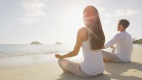 Young couple meditating together in lotus position on shore at beach. Relaxed man and woman are in sports clothing. They are performing yoga in front of sea during sunny day.