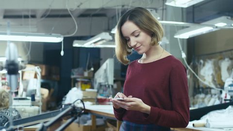 Young woman is standing in a clothing factory and using a smartphone while employees work in the background. Shot on RED Cinema Camera in 4K (UHD)