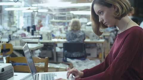 Young female is sitting at a table in a clothing factory and working on a laptop while employees are sewing in the background. Shot on RED Cinema Camera in 4K (UHD)