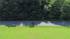 A small red vintage car is passing a forest while driving on a country rouad. Wide-angle shot from the side. There is also a green meadow seen in the front of the video. The weather is gorgeous.
