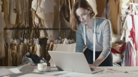 Woman clothing designer is working on a laptop while leaning on a studio table. Shot on RED Cinema Camera in 4K (UHD).