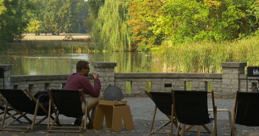 Man sits relaxed in the deckchair. He lays back in the chair and drinks from the cup in his hand. Man in maroon shirt and beige pants. Back view. Park area, lake, green trees, dry yellow leaves.
