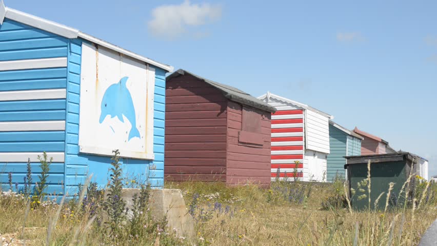 Row of wooden beach huts on a sunny but windy day, with the grass in the foreground blowing in the breeze, the sky is clear and blue.