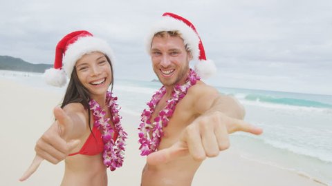 Christmas Hawaii vacation - Hawaiian beach couple wearing santa hat and doing welcome shaka sign happy at camera as welcoming gesture for winter holidays.