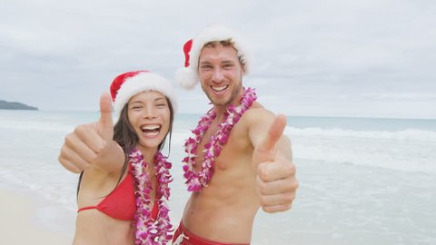 Christmas beach Hawaii vacation couple happy giving thumbs up. Hawaiian beach couple wearing santa hat and Lei flower garland happy at camera on winter holidays. RED EPIC SLOW MOTION.