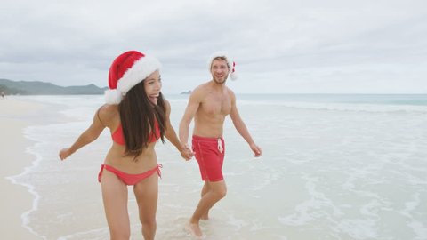 Christmas couple happy relaxing on white sand beach running on sand in bikini and swimsuit. Asian woman and man holding hands running playing during travel holidays. RED EPIC SLOW MOTION.