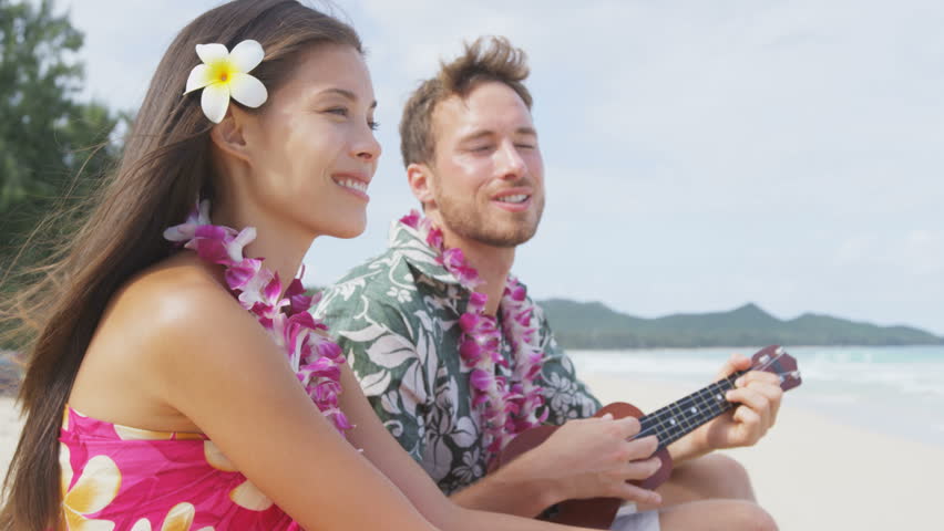 Man on beach playing ukulele instrument on Hawaii with having fun. Young couple, woman and man in love on beach vacations in Hawaiian clothing wearing Aloha shirt dress and flower lei. RED EPIC.