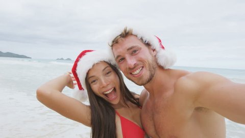 Christmas couple selfie picture on beach vacation. Happy young adults smiling at camera taking self-portrait wearing santa hats. Multiracial Caucasian and Asian people. RED EPIC SLOW MOTION.