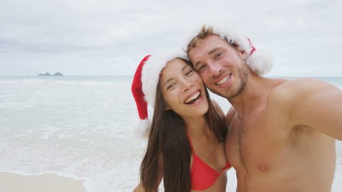 Happy couple on Christmas travel holidays taking selfie picture with smartphone wearing santa hat during their winter vacation. Young adult friends in swimsuit and bikini in by the ocean. RED EPIC.