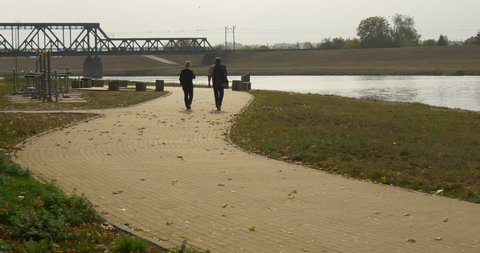 Man and woman walk on the road paved with paving slabs. Man threw a jacket over his shoulder. They walk near the sports ground in the park. Bridge across the small river on the background.