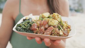 Midsection of young female holding poke salad plate. Closeup of woman with traditional Hawaii dish consisting of raw marinated ahi yellowfin tuna fish. Tourist is sitting at beach.