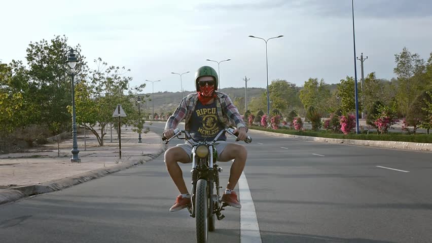 Young happy man in helmet riding motorbike on highway road on green trees background. Slow motion