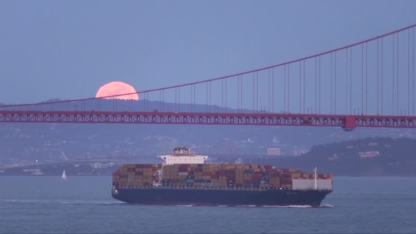 Tanker disembarking the San Francisco Bay under the Golden Gate Bridge with the light of a rising orange full moon over the Berkeley Hills behind.