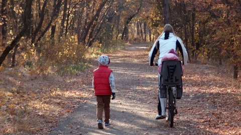 Mother rides the daughter on the bicycle in autumn park and the son runs nearby 