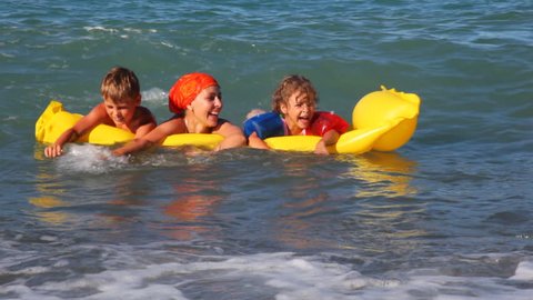 mother with two kids swim in wavy sea, holding yellow inflatable mattress sunny day