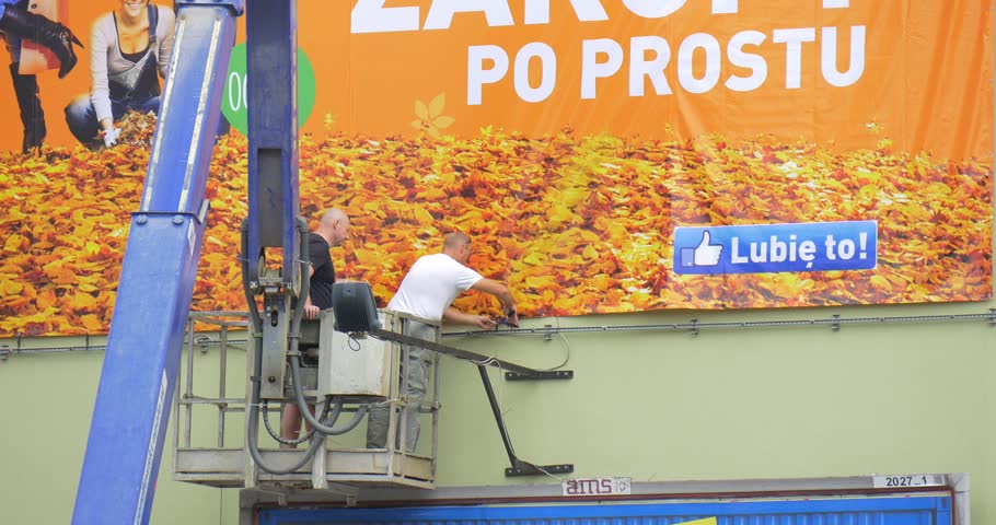 Two Men Workers in Truck Tower are Installing an Orange Advertisement Banner into Metal Frame to the building, Tower Moves Up, Moves Down, Moves to the Right, city, people are working outdoors, fall,