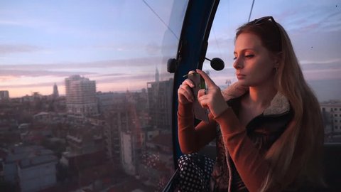 Travel: beautiful young woman tourist looking through the glass, making photos with her cellphone in the ropeway cabin at sunset. Medium shot, slow motion 60 fps, handheld, HD.