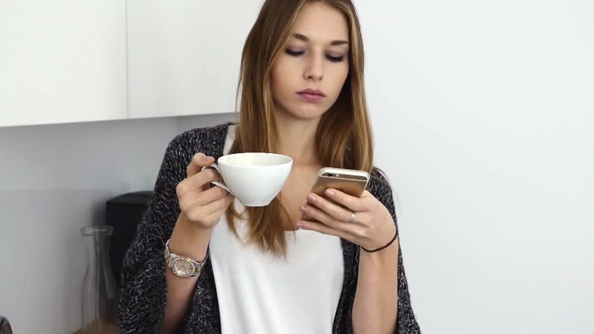 Beautiful young girl drinkin coffee and using smartphone in the kitchen. Indoor photo.