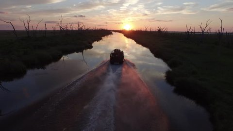 An airboat powers down a waterway during a beautiful southern Louisiana sunset over a bayou and stretch of water dotted with silhouettes of dead trees.