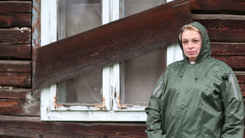 Woman in waterproof raincoat stands near the boarded-up uninhabited house and looks at the camera