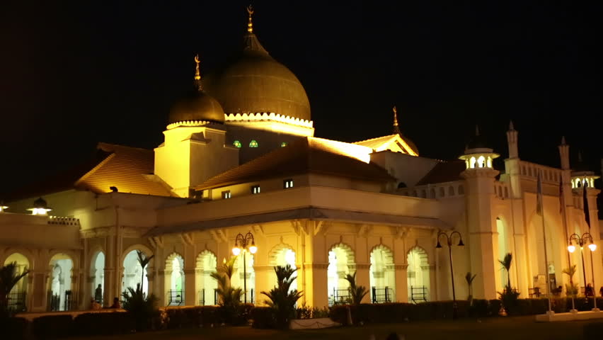 External night illuminated Kapitan Keling Mosque (Penang) with dome and white walls, people leaving and entering prayer area, black night sky