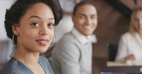 Portrait of confident hispanic business woman at boardroom table in trendy modern shared office space a diverse team chatting together in background In slow motion turning around and smiling