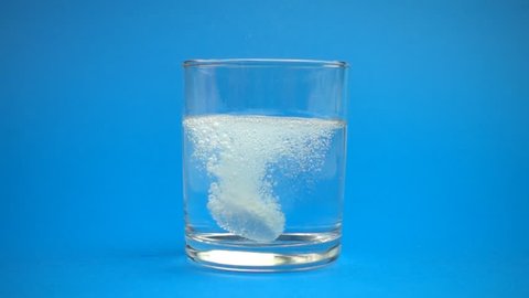 Aspirin or effervescent pill dropping into a glass of water slow motion on blue background.