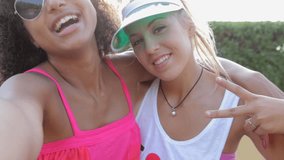 Portrait of two ethnically diverse teenager girls friends holding heads together pulling faces at the camera while taking a selfie video of themselves, outdoors lifestyle. Americana colorful vintage.