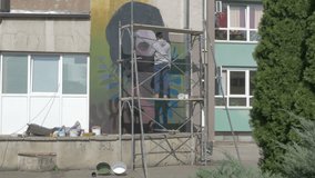Talented young graffiti artist standing on scaffold and painting mural with brush. Performance,raw video,no grading. Man drawing graffiti on wall with spray bottle. Male painter performing street art.
