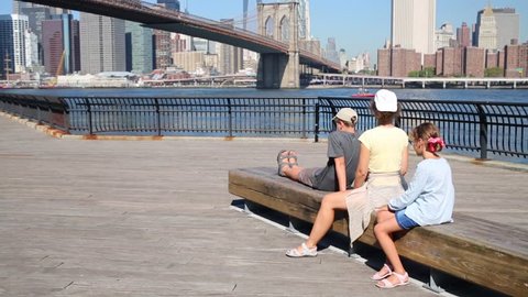 Family of three sit on bench on quay near Brooklyn Bridge in New York city
