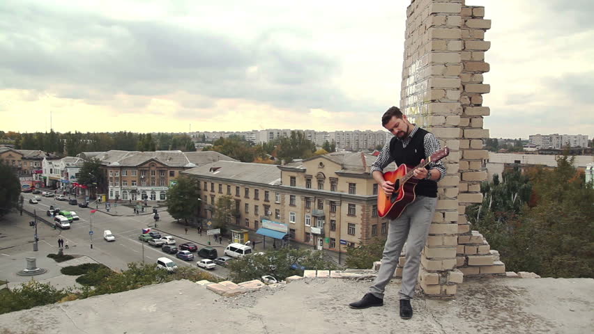 The roof of an unfinished house, against the backdrop of a provincial town. A man play on the guitar.