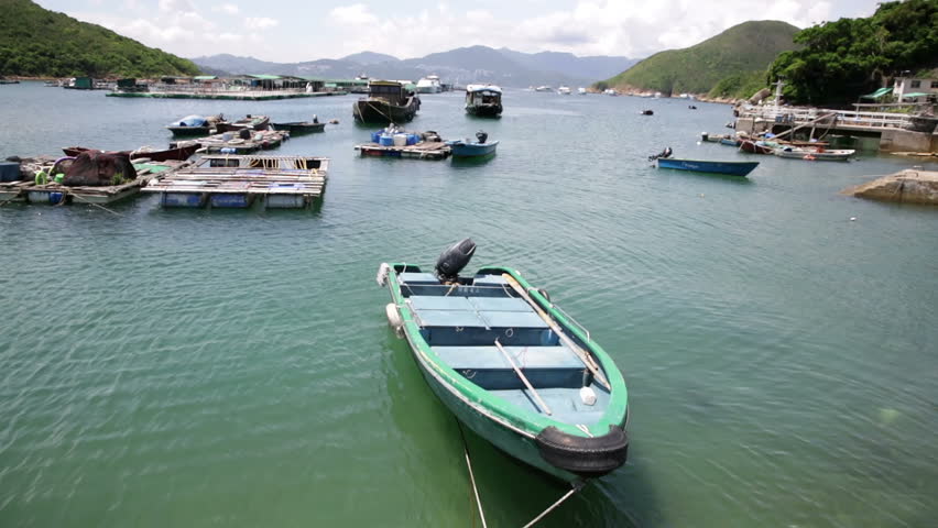 Small boat tied up with other boats visible at harbour in Sai Kung area of Hong Kong, blue emerald sea floating fish containers, hills and sky on a late Summer sunny hot day.