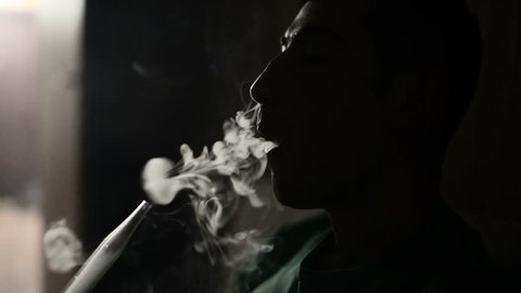 Young man smokes hookah and lets out smoke rings in a dark room close up.