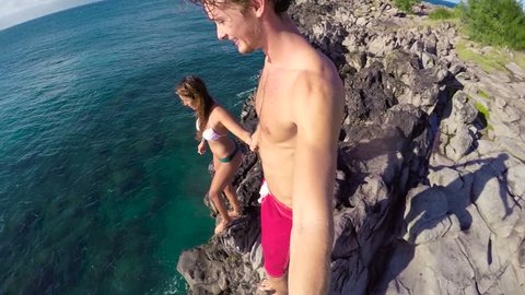 POV Slow Motion Cliff Jumping. Athletic Young Couple Holding Hands Jumping From Cliff Into Ocean. Adventure Extreme Sports Lifestyle Hobby Vacation