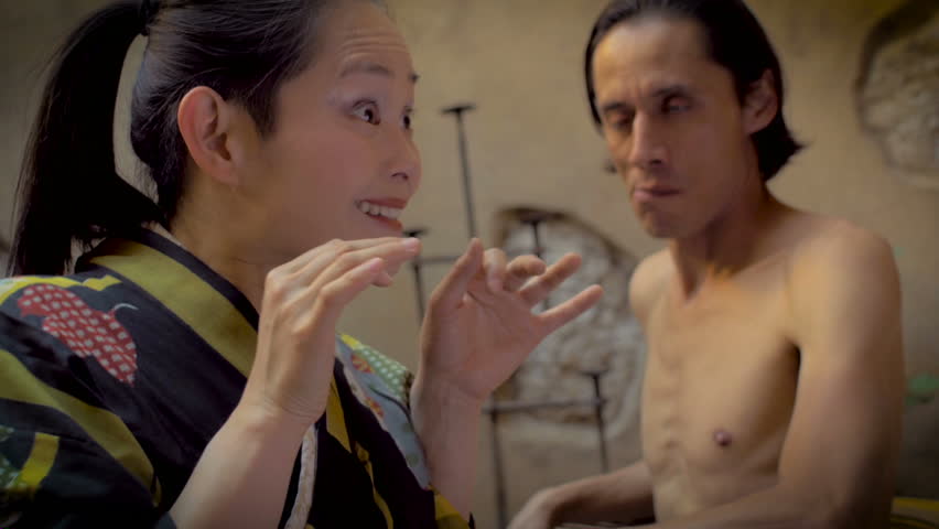 Dolly shot of a Japanese woman screaming with her hands on her face while a man without a shirt reacts because her yelling is hurting his ears. Both are performing a modern dance routine.