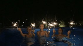 Slow motion clip of two happy women and a man dancing with sparklers in the swimming pool at night. Christmas and New Year celebration