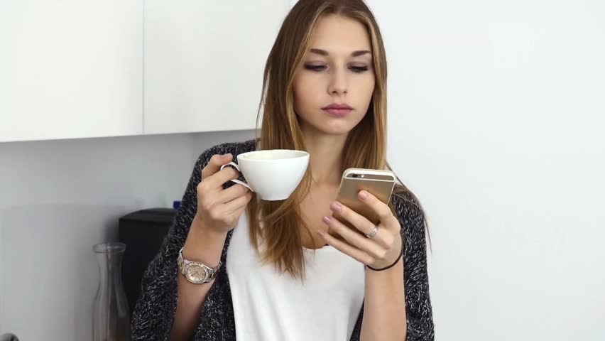 Beautiful young girl drinkin coffee and using smartphone in the kitchen. Indoor photo.