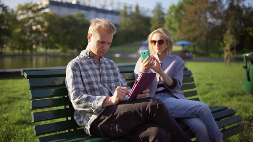 Couple sitting on the bench in the park and using electronics
