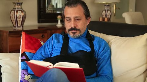 Painful man with injured arm and bandage sitting on sofa and reading a book