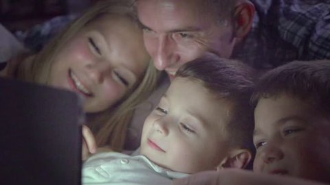 Happy family- Father, mother and Two kids lying down on the sofa and using tablet pc at night. Family watching movie on tablet computer in a dark room. HD 1080p, slow motion 240 fps, high speed camera