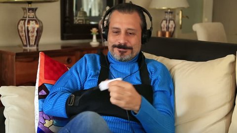 Painful man with a broken arm wearing arm brace sitting on a sofa and listening to music