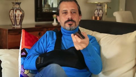 Painful man with a broken arm wearing arm brace sitting on sofa and watching tv